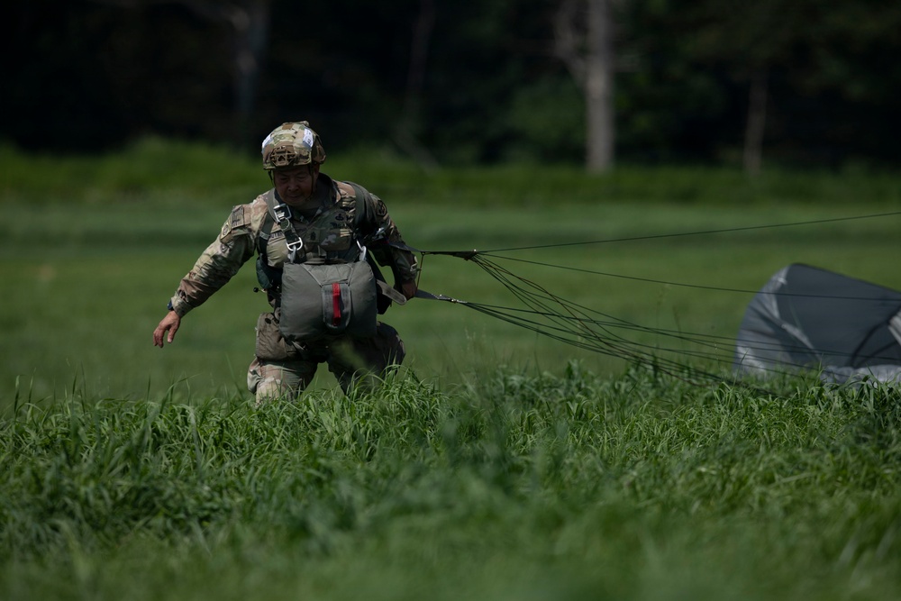 A military paratrooper in full camouflage uniform and helmet is walking through a grassy field after a parachute landing. The soldier is dragging a collapsed parachute behind them, with the canopy and suspension lines visible on the ground to the right. The setting appears to be a designated drop zone surrounded by trees in the background.