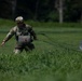 A military paratrooper in full camouflage uniform and helmet is walking through a grassy field after a parachute landing. The soldier is dragging a collapsed parachute behind them, with the canopy and suspension lines visible on the ground to the right. The setting appears to be a designated drop zone surrounded by trees in the background.