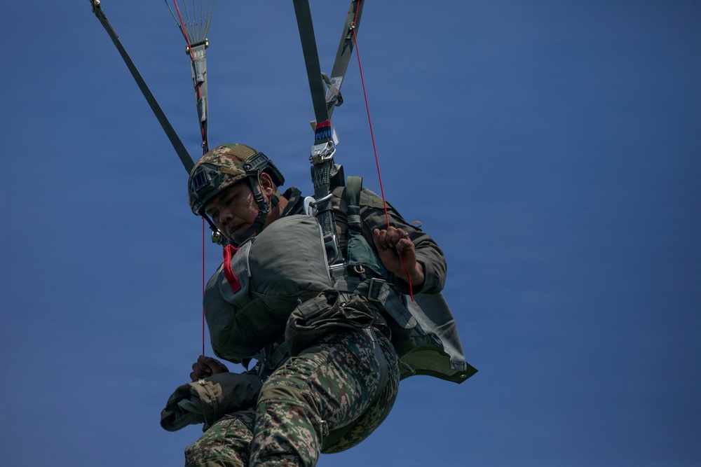 A military paratrooper is suspended in mid-air beneath a parachute canopy against a clear blue sky. The soldier is wearing full combat gear including a camouflage uniform, tactical helmet with goggles, body armor, and a harness system with visible red control lines. The image is taken from a low angle looking upward, capturing the soldier during a parachute descent or training exercise.