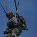 A military paratrooper is suspended in mid-air beneath a parachute canopy against a clear blue sky. The soldier is wearing full combat gear including a camouflage uniform, tactical helmet with goggles, body armor, and a harness system with visible red control lines. The image is taken from a low angle looking upward, capturing the soldier during a parachute descent or training exercise.