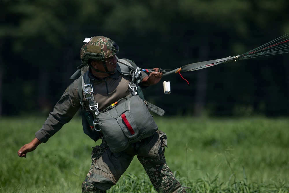 A military paratrooper is shown on a grassy field immediately after landing, gathering parachute lines and canopy. The soldier is wearing full combat gear including a camouflage helmet, parachute harness, and tactical uniform. The image captures the post-landing recovery phase of a parachute operation.