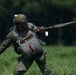 A military paratrooper is shown on a grassy field immediately after landing, gathering parachute lines and canopy. The soldier is wearing full combat gear including a camouflage helmet, parachute harness, and tactical uniform. The image captures the post-landing recovery phase of a parachute operation.