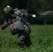 A military paratrooper is shown on the ground in a green grassy field after a parachute landing. The soldier is wearing digital camouflage uniform and helmet while managing parachute lines and risers. The image captures the immediate post-landing phase as the trooper works to control the deployed parachute canopy lines.