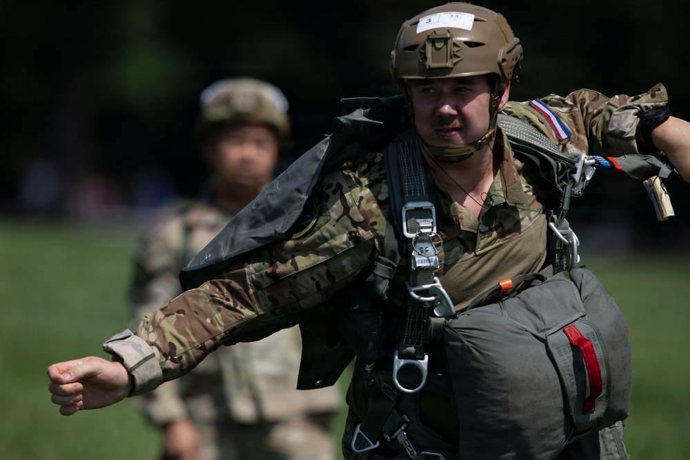 A military paratrooper in full combat gear and parachute harness is walking in an outdoor area, appearing to be preparing for or returning from a jump exercise. The soldier wears a tan tactical helmet with a number label, multicam camouflage uniform, and carries a packed parachute on their back. A second soldier in similar attire is visible but out of focus in the background.