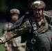 A military paratrooper in full combat gear and parachute harness is walking in an outdoor area, appearing to be preparing for or returning from a jump exercise. The soldier wears a tan tactical helmet with a number label, multicam camouflage uniform, and carries a packed parachute on their back. A second soldier in similar attire is visible but out of focus in the background.