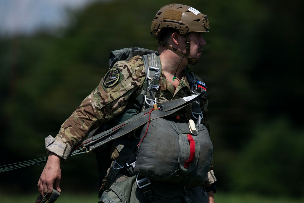 A military paratrooper is pictured in profile view wearing full jump gear including a parachute harness and reserve chute. The soldier is dressed in MultiCam (OCP) camouflage uniform with a tan ballistic helmet and a unit patch visible on the left shoulder. The image appears to be taken on a grassy drop zone or staging area with a blurred green tree line in the background.