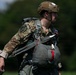 A military paratrooper is pictured in profile view wearing full jump gear including a parachute harness and reserve chute. The soldier is dressed in MultiCam (OCP) camouflage uniform with a tan ballistic helmet and a unit patch visible on the left shoulder. The image appears to be taken on a grassy drop zone or staging area with a blurred green tree line in the background.