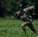 A military paratrooper in multicam camouflage uniform has just landed in a grassy field and is running forward while managing parachute lines trailing behind him. He is wearing a black helmet and full parachute harness with equipment. A flag patch visible on his right shoulder appears to be the French tricolor (blue, white, red).