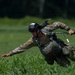 A military paratrooper is captured mid-landing in a green grassy field, having just touched down from a parachute jump. The soldier is in a dynamic, off-balance posture, holding parachute control toggles/risers in one hand while extending the other arm for balance. The image is a high-quality action shot with sharp focus on the subject against a blurred natural background.