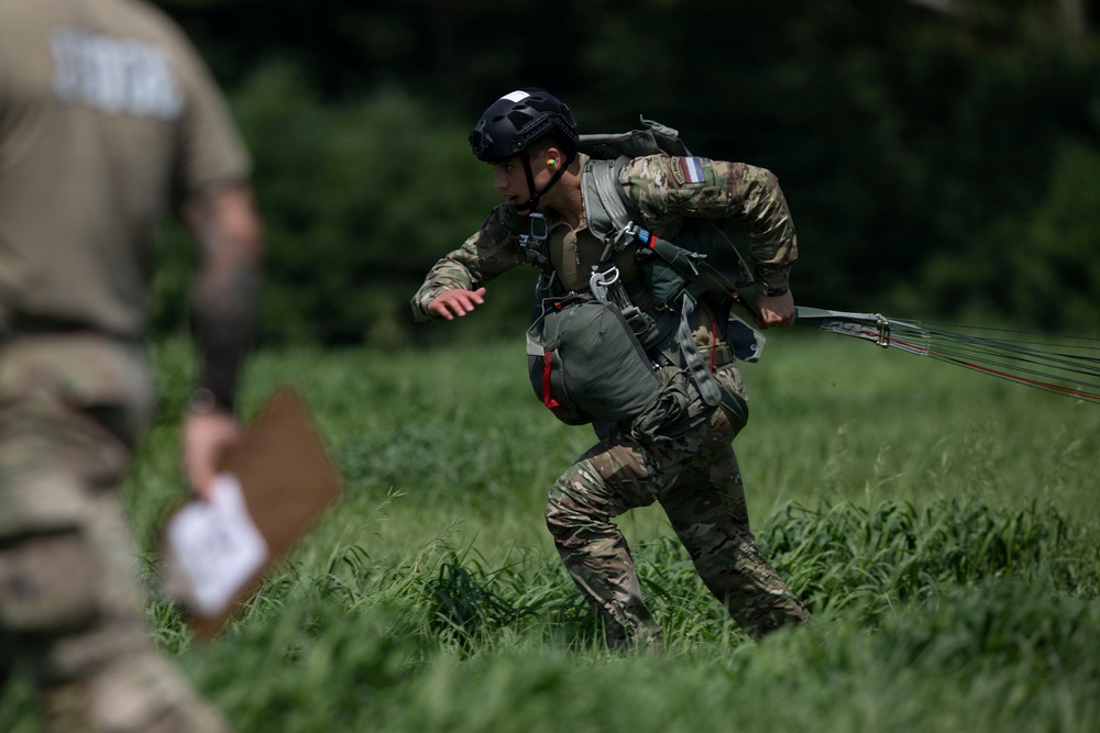 A military paratrooper in full combat gear is moving across a grassy field after an apparent parachute landing, with parachute lines still trailing behind them. The soldier is wearing multicam camouflage, a tactical helmet, and carrying a reserve parachute pack. A second individual in the foreground, partially blurred, appears to be an observer or judge holding a clipboard.