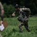 A military paratrooper in full combat gear is moving across a grassy field after an apparent parachute landing, with parachute lines still trailing behind them. The soldier is wearing multicam camouflage, a tactical helmet, and carrying a reserve parachute pack. A second individual in the foreground, partially blurred, appears to be an observer or judge holding a clipboard.