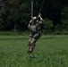 A military paratrooper is captured moments before or during landing in a green grassy field. The soldier is in full combat gear, suspended by parachute risers and toggle lines, appearing to be steering or preparing for touchdown. The background features dense, dark green forest trees, suggesting a rural or training drop zone.