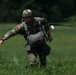 A U.S. Army soldier in full parachute gear is moving across a grassy field after an airborne landing. The soldier is wearing MultiCam (OCP) camouflage uniform with a helmet, parachute harness, and reserve parachute, with parachute suspension lines trailing behind. The setting appears to be a designated drop zone during a military airborne training or operational jump.