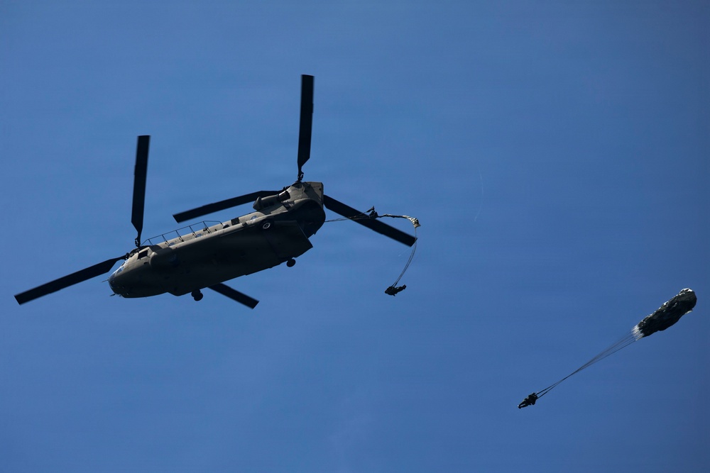 A large tandem-rotor military helicopter, consistent with a CH-47 Chinook, is photographed from below against a clear blue sky. At least two paratroopers are visible in the process of exiting the aircraft, with one parachute already deployed to the right of the helicopter. This appears to be a military airborne operation or training exercise.