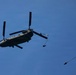 A large tandem-rotor military helicopter, consistent with a CH-47 Chinook, is photographed from below against a clear blue sky. At least two paratroopers are visible in the process of exiting the aircraft, with one parachute already deployed to the right of the helicopter. This appears to be a military airborne operation or training exercise.