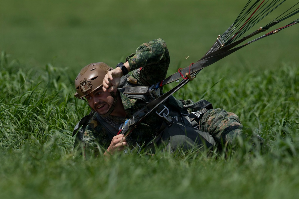 A military paratrooper has just landed in a grassy field and is lying on the ground managing his parachute lines. He is wearing camouflage uniform, a tan/brown tactical helmet, and full parachute harness. The soldier appears to be in the process of collapsing or releasing his parachute canopy after landing.