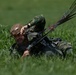 A military paratrooper has just landed in a grassy field and is lying on the ground managing his parachute lines. He is wearing camouflage uniform, a tan/brown tactical helmet, and full parachute harness. The soldier appears to be in the process of collapsing or releasing his parachute canopy after landing.