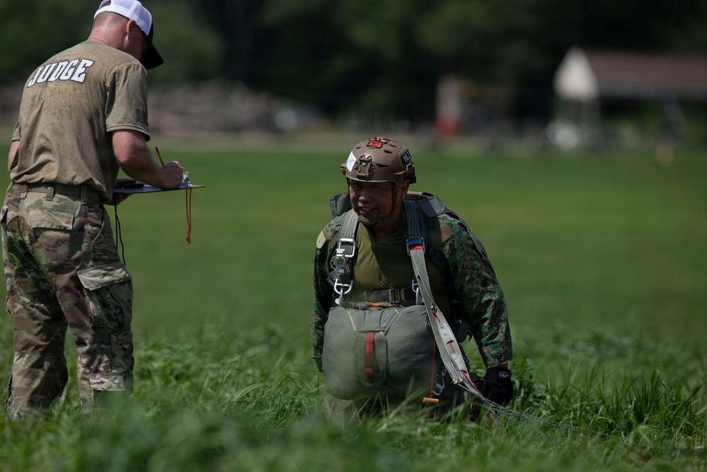 Two military personnel are visible in an open grassy field. One individual wearing a 'JUDGE' t-shirt and camouflage pants is holding a clipboard and appears to be recording information. The second individual is wearing full parachute gear including a tactical helmet, harness, and parachute pack, suggesting this is a post-jump landing zone assessment.
