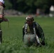 Two military personnel are visible in an open grassy field. One individual wearing a 'JUDGE' t-shirt and camouflage pants is holding a clipboard and appears to be recording information. The second individual is wearing full parachute gear including a tactical helmet, harness, and parachute pack, suggesting this is a post-jump landing zone assessment.