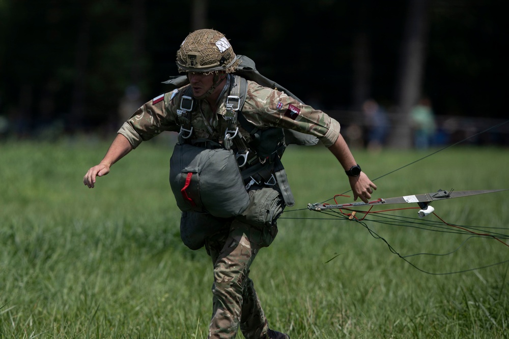 A military paratrooper in full combat gear is running across a grassy field immediately after landing, still holding parachute lines in his right hand. The soldier is wearing British MTP (Multi-Terrain Pattern) camouflage uniform with a netted helmet, parachute harness, and a leg equipment bag. Patches on the uniform suggest British military affiliation, possibly during a commemorative or training jump event.