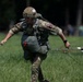 A military paratrooper in full combat gear is running across a grassy field immediately after landing, still holding parachute lines in his right hand. The soldier is wearing British MTP (Multi-Terrain Pattern) camouflage uniform with a netted helmet, parachute harness, and a leg equipment bag. Patches on the uniform suggest British military affiliation, possibly during a commemorative or training jump event.