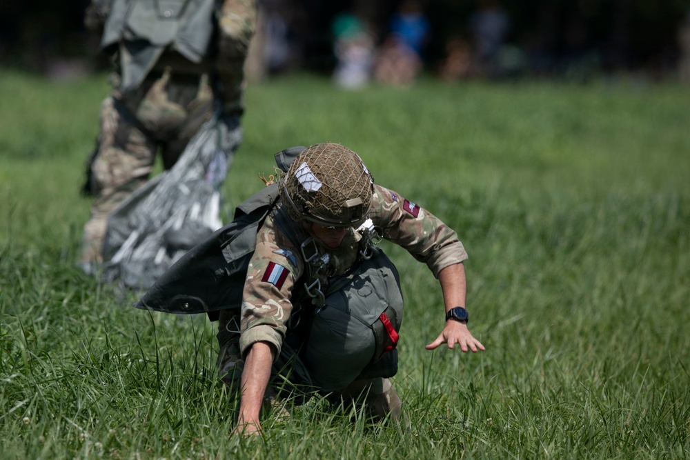 A military paratrooper has just landed on a grassy field and is in the process of recovering from touchdown, crouching low to the ground with hands bracing against the grass. The soldier is wearing British military multicam camouflage uniform with a netted helmet, parachute harness, and what appears to be a maroon and blue patch on the arm, consistent with British Parachute Regiment insignia. A second soldier and a partially visible parachute canopy can be seen in the background.