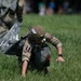 A military paratrooper has just landed on a grassy field and is in the process of recovering from touchdown, crouching low to the ground with hands bracing against the grass. The soldier is wearing British military multicam camouflage uniform with a netted helmet, parachute harness, and what appears to be a maroon and blue patch on the arm, consistent with British Parachute Regiment insignia. A second soldier and a partially visible parachute canopy can be seen in the background.