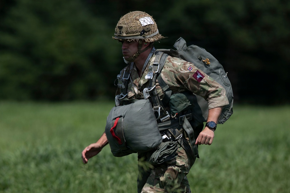 A soldier in British military camouflage uniform is moving across a grassy field while carrying full parachute equipment and a large pack. The soldier wears a netted helmet with markings and has multiple harness straps and gear attached to their body. The image appears to depict a post-jump recovery scenario, likely during a military airborne exercise.