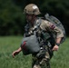 A soldier in British military camouflage uniform is moving across a grassy field while carrying full parachute equipment and a large pack. The soldier wears a netted helmet with markings and has multiple harness straps and gear attached to their body. The image appears to depict a post-jump recovery scenario, likely during a military airborne exercise.