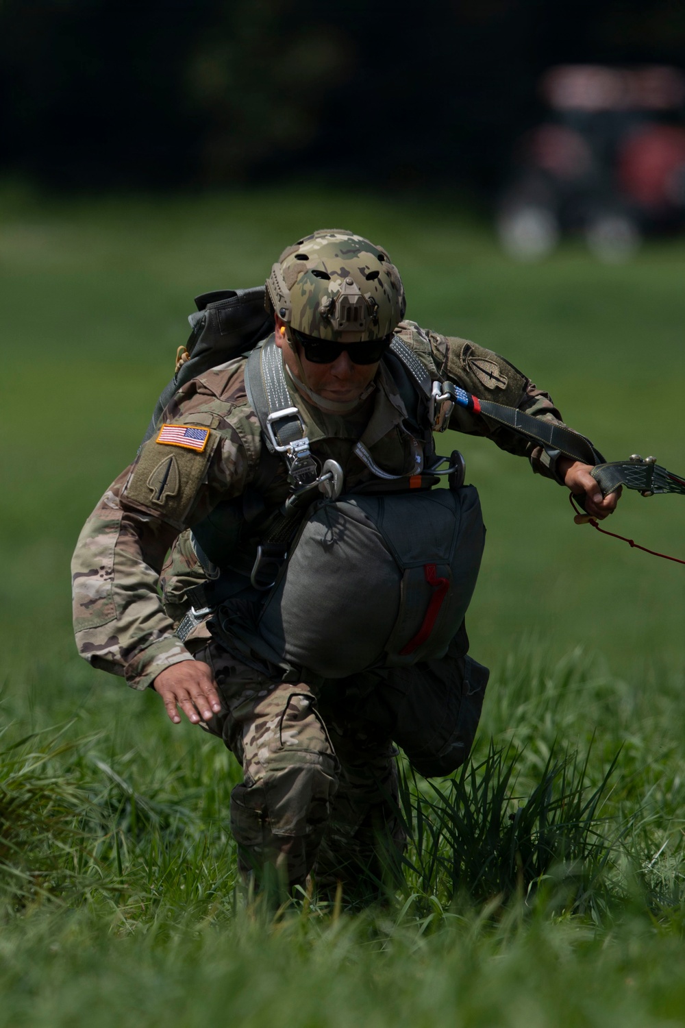 A U.S. Army paratrooper is captured on a grassy landing zone immediately after a parachute landing, crouching low while gathering parachute lines. The soldier is wearing full multicam combat uniform with a tactical helmet, parachute harness, and reserve parachute container. The American flag patch is clearly visible on the left shoulder, identifying this as a U.S. military operation.