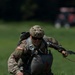 A U.S. Army paratrooper is captured on a grassy landing zone immediately after a parachute landing, crouching low while gathering parachute lines. The soldier is wearing full multicam combat uniform with a tactical helmet, parachute harness, and reserve parachute container. The American flag patch is clearly visible on the left shoulder, identifying this as a U.S. military operation.