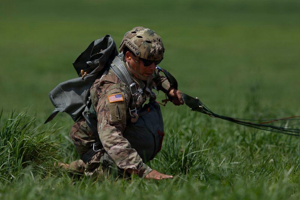 A U.S. Army soldier has just landed in a grassy field following a parachute jump, identifiable by the parachute lines and canopy visible trailing behind them. The soldier is wearing full multicam combat uniform with a tactical helmet, sunglasses, parachute harness, and reserve parachute pack. They appear to be in the process of recovering from landing, crouching low and gathering parachute lines.
