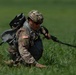 A U.S. Army soldier has just landed in a grassy field following a parachute jump, identifiable by the parachute lines and canopy visible trailing behind them. The soldier is wearing full multicam combat uniform with a tactical helmet, sunglasses, parachute harness, and reserve parachute pack. They appear to be in the process of recovering from landing, crouching low and gathering parachute lines.