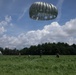 A military parachute operation is underway on a green open field surrounded by dense tree line. A large olive/silver round parachute canopy is fully deployed and descending, while several soldiers in camouflage uniforms are on the ground managing equipment. A second collapsed parachute is visible to the left, and what appears to be an aircraft is faintly visible in the upper right sky.