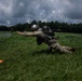 A military paratrooper in camouflage uniform and helmet is kneeling on a grassy field, appearing to have just landed after a parachute jump. The soldier is still connected to parachute lines and is in the process of recovering from landing. The scene takes place on an open drop zone with overcast skies and tree lines visible in the background.