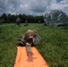 A military airborne operation is depicted on a grassy drop zone, with multiple soldiers having recently landed via parachute. A foreground soldier in full combat gear is prone on an orange ground marker panel, while a large olive-drab parachute is still inflated in the background. Multiple other soldiers in U.S. Army camouflage uniforms are moving across the field post-landing.