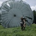 A military paratrooper has just landed in a green grassy field, with a large gray/olive-colored round parachute still inflated and billowing behind them. The soldier is wearing full combat gear including camouflage uniform, helmet, and harness with equipment attached. The setting appears to be a training drop zone surrounded by trees under a partly cloudy sky.