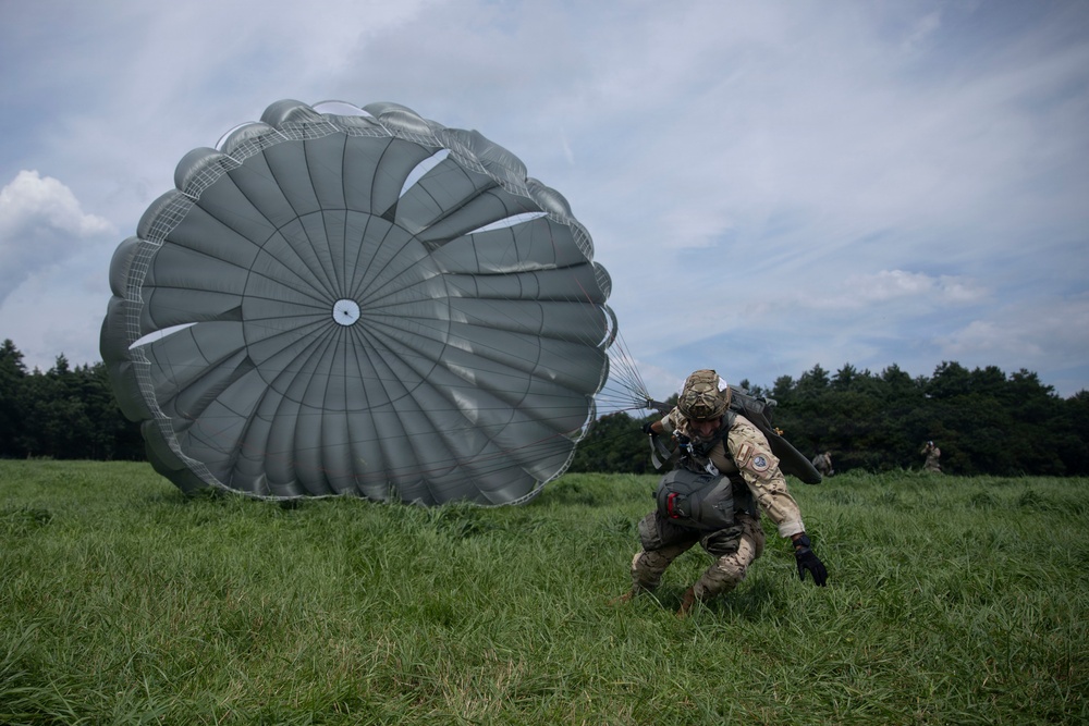A military paratrooper has just landed in a grassy field and is being dragged forward by a large gray round parachute still inflated by wind. The soldier is in full combat gear and is attempting to collapse or control the parachute canopy. Additional personnel are visible in the background, suggesting a group airborne operation or training exercise.