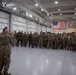 A military briefing or address is taking place inside a large hangar facility, where a single uniformed soldier stands with their back to the camera addressing a large assembled group of troops. An American flag is prominently displayed on the back wall, and multiple national or unit flags are visible in the background. The gathering appears to be a pre-deployment briefing, unit formation, or official address involving a multinational or multi-unit military assembly.