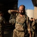 Military personnel are gathered near a large truck or trailer in what appears to be a loading dock or staging area. A bearded, bald soldier in MultiCam/MTP camouflage uniform with a harness is prominently featured in the foreground, adjusting equipment on his back. In the background, another soldier wearing a 'RIGGER' t-shirt with parachutist wings insignia is working at the truck, while additional uniformed personnel are visible to the right.