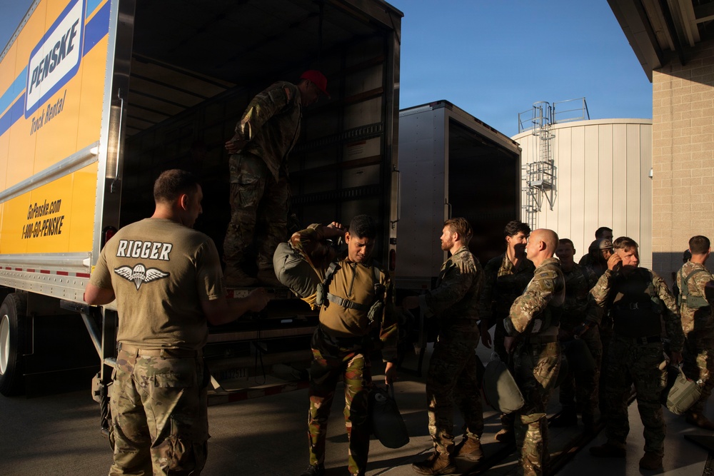 A group of military personnel in camouflage uniforms are loading or unloading gear from a yellow Penske rental truck. One soldier wearing a 'RIGGER' t-shirt with parachutist wings stands near the truck's rear, while another soldier is inside the truck bed. The scene appears to take place at a military installation or staging area during daylight hours.