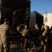 A group of military personnel in camouflage uniforms are loading or unloading gear from a yellow Penske rental truck. One soldier wearing a 'RIGGER' t-shirt with parachutist wings stands near the truck's rear, while another soldier is inside the truck bed. The scene appears to take place at a military installation or staging area during daylight hours.
