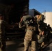 Military personnel in camouflage uniforms are loading or unloading equipment from a yellow Penske rental truck. A soldier with 'RIGGER' printed on the back of their uniform and parachutist wings insignia is prominently visible, suggesting airborne/parachute unit involvement. The scene appears to be a logistics or deployment operation taking place at a building loading dock.