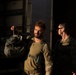 The image shows military personnel in tactical gear near the open rear of a large vehicle or container. The primary subject, a bearded male in multicam/OCP uniform with a plate carrier vest, is holding what appears to be a tactical helmet. The scene is dramatically lit with strong directional sunlight creating high contrast shadows.