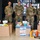 Four military personnel in OCP (Operational Camouflage Pattern) uniforms are posing in front of a 'Welcome to' sign at what appears to be a military installation. They are standing behind a collection of donated goods including hygiene products, laundry detergent, and care package items arranged in cardboard boxes. The setting suggests a charitable donation drive or care package assembly event.