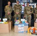 Four military personnel in OCP (Operational Camouflage Pattern) uniforms are posing in front of a 'Welcome to' sign at what appears to be a military installation. They are standing behind a collection of donated goods including hygiene products, laundry detergent, and care package items arranged in cardboard boxes. The setting suggests a charitable donation drive or care package assembly event.