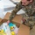 A U.S. Air Force service member, identified by name tape as 'ADAMS' (or similar), is kneeling on a tiled floor and sorting through or packing a large cardboard box filled with food and supply items. The individual is wearing Operational Camouflage Pattern (OCP) uniform with U.S. Air Force branch tape clearly visible. This appears to be a food distribution, care package assembly, or supply logistics activity.
