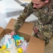 A U.S. Air Force service member, identified by name tape as 'ADAMS' (or similar), is kneeling on a tiled floor and sorting through or packing a large cardboard box filled with food and supply items. The individual is wearing Operational Camouflage Pattern (OCP) uniform with U.S. Air Force branch tape clearly visible. This appears to be a food distribution, care package assembly, or supply logistics activity.