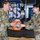 Two U.S. Air Force personnel in OCP (Operational Camouflage Pattern) uniforms are exchanging or presenting a large orange container of Tide Pods laundry detergent. They are standing in front of a sign that reads 'Welcome to [something] GS-1' with what appears to be a military unit shield/emblem. Both individuals are smiling, suggesting a lighthearted or ceremonial moment.
