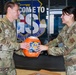 Two U.S. Air Force personnel in OCP (Operational Camouflage Pattern) uniforms are exchanging or presenting a large orange container of Tide Pods laundry detergent. They are standing in front of a sign that reads 'Welcome to [something] GS-1' with what appears to be a military unit shield/emblem. Both individuals are smiling, suggesting a lighthearted or ceremonial moment.